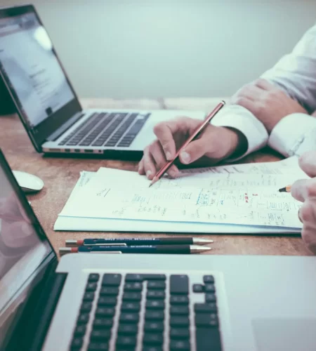 Twee mensen werken samen aan een bureau met open laptops en papieren met notities.